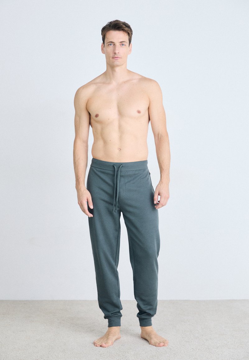 Dunkelgraue Herren-Sweatpants mit Kordelzugbund; aus weichem Material gefertigt, mit schmal zulaufenden Beinen und gerippten Bündchen, barfuß getragen.