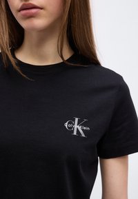 Νεαρή γυναίκα που φοράει μαύρο t-shirt Calvin Klein με μικρό λευκό "CK" και το όνομα της μάρκας τυπωμένα στο αριστερό μέρος του στήθους.