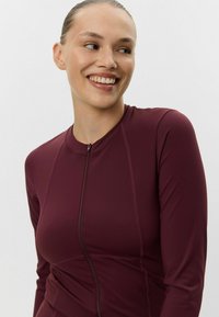 Vein punane pikkade varrukatega topp, millel on tõmblukk, sobituv disain ja sile tekstuur. Kaunistatud õmbluse detailidega.