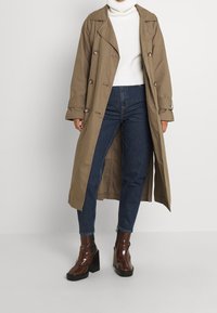 Kaki trenchcoat med dubbelknäppta knappar, bred krage och sidofickor, kombinerat med en vit polotröja och mörkblå jeans, accentuerad med bruna klackstövlar.