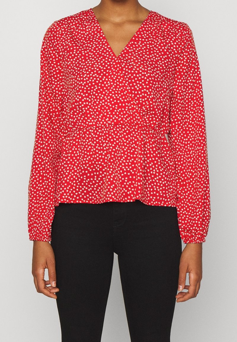 Blouse rouge avec un motif de cœurs blancs, décolleté en V, manches longues et taille froncée. Associée à un pantalon noir.