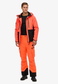 Felroze waterdichte jas en broek met zwarte accenten en cargozakken. Textuurstof, capuchonontwerp en ritsclosures.