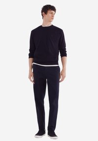 Marinefarbener Pullover mit Rundhalsausschnitt, getragen über einem weißen Hemd. Dazu navyfarbene Hosen und schwarze Sneakers, die ein minimalistisches Design aufweisen.