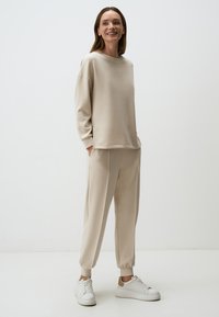 Beige fleece sweatshirt met lange mouwen en een ronde hals, gecombineerd met aansluitende tapered broek. Voorzien van elastische boorden en witte sneakers.