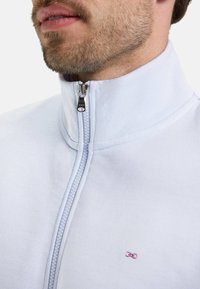 Veste zippée bleu clair avec un col montant, dotée d'une fermeture éclair à l'avant et d'un petit logo rose sur la poitrine. Texture de tissu lisse.