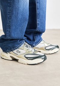 Buty sportowe Lacoste mają cholewkę z siateczki i materiału syntetycznego w kolorach białym, srebrnym i ciemnozielonym. Gruba gumowa podeszwa z teksturowanymi akcentami oraz sznurowadłami.