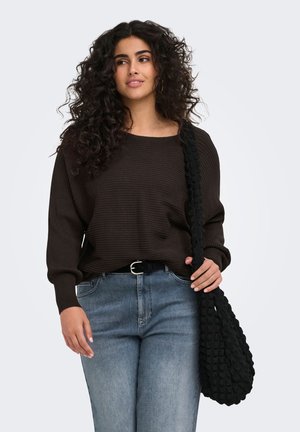 Kvinde med krøllet hår iført en mørk ribstrikket sweater og blå jeans, der bærer en stor tekstureret sort skuldertaske.