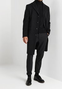 Cappotto in lana nera con un design strutturato, chiusura a bottoni e due tasche frontali; indossato sopra un dolcevita nero e pantaloni su misura.