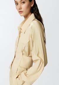 Bež satenasti kratki sako s oštrim ovratnikom, prednjim gumbima i napuhanim rukavima; ima strukturirani kroj i bočnu džep.