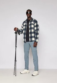 Άνδρας που φοράει μαύρο και άσπρο καρό σακάκι, μπλε παντελόνια, λευκά αθλητικά παπούτσια, γυαλιά ηλίου, κρατώντας μαύρα μπατόν πεζοπορίας σε απλό φόντο.