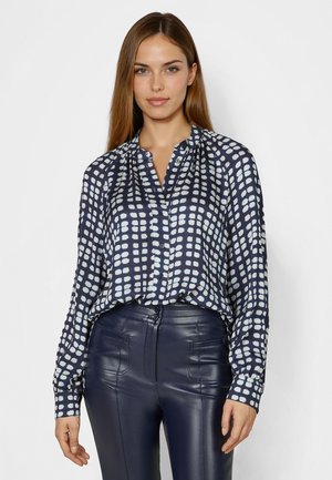 Woman wearing navy blue and white checkered long-sleeve blouse tucked into high-waisted navy blue leather pants, standing against a plain background.