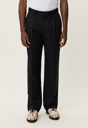 Pantaloni neri a vita alta con pieghe frontali, realizzati in tessuto dalla trama liscia, abbinati a scarpe bianche e marroni con motivo decorativo.