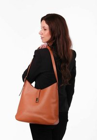 Hnědá kožená kabelka přes rameno s uvolněným tvarem, dlouhým ramenním popruhem a jemným metalickým akcentem na přední straně.