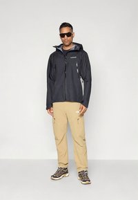 Black waterproof jacket with zippered pockets, beige cargo pants, and rugged hiking shoes. Minimalist design, suitable for outdoor activities.