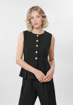 Kvinne med skulderlangt blond hår som har på seg en ermeløs svart topp med fem gullknapper, stående foran en ensfarget lys bakgrunn.