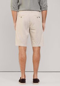 Beige katoenen shorts met een eenvoudig ontwerp, voorzien van twee achterzakken en knopen, omgeslagen zoom en gedragen met donkere schoenen.