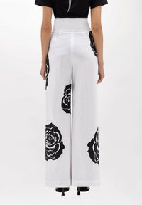 Pantaloni bianchi a gamba larga con grandi motivi di rose nere. Realizzati in un tessuto leggero con una vita alta. Calzature nere visibili.