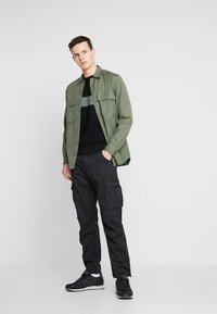 Grønne knappellede skjorte, sort langærmet sweater med grøn stribe, sorte cargo bukser og sorte sneakers. Casual, lagdelt outfit.
