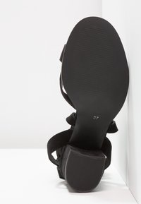 Sandale noire à talons hauts avec une tige en velours texturé, une sangle ajustable et une semelle en caoutchouc lisse. Taille 37 indiquée sur la semelle.