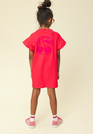 Meisje met haar in een knot, draagt een rode bril en een rode jurk met kersenmotief en de tekst "Let's Hang Out", roze sneakers, staat met de rug naar ons toe.