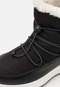 Stivale waterproof nero con fodera in pelliccia sintetica, sistema di allacciatura elastico, tessuto testurizzato e suola in gomma bianca. Superiori lisce e imbottite.
