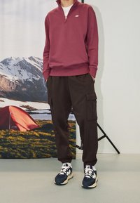 Bordeaux zip-up sweatshirt, bruine cargo broek met zakken, en marineblauwe sneakers met witte accenten, tegen een natuurachtergrond.