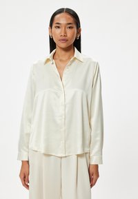 Blusa de satén color crema con cuello, cierre de botones en el frente y mangas largas. Textura suave y corte relajado.