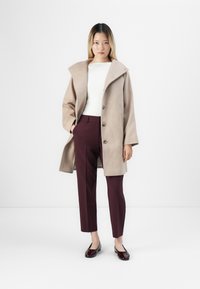 Femme jeune debout portant un manteau en laine beige, un haut blanc, un pantalon bordeaux et des ballerines rouge foncé sur un fond blanc uni.