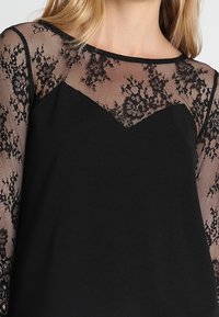 Top nero con una parte superiore in pizzo trasparente con motivi floreali e maniche lunghe. Il collo presenta una delicata scollatura a V.