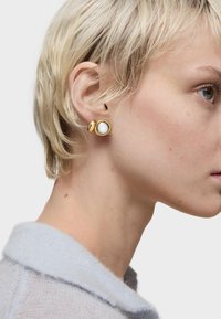 Orecchini in tono oro con una pietra bianca rotonda in una montatura circolare, abbinati a un piccolo accento in oro lucido, indossati da un modello con capelli corti.