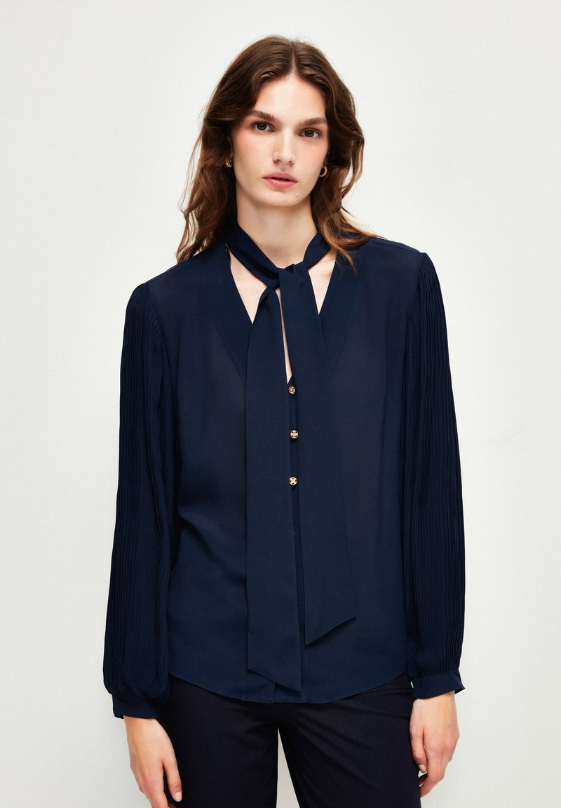 Blusa blu navy con colletto a fiocco, maniche lunghe plissettate e bottoni dorati sul davanti. Il tessuto appare leggero e dalla texture liscia.