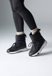 Bottes noires imperméables avec un dessus matelassé, des détails de laçage et une semelle en caoutchouc blanche. Portées avec des leggings noirs et des chaussettes de couleur claire.