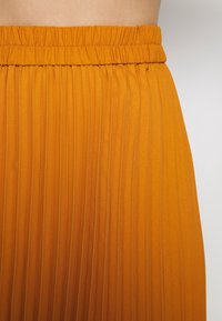 Jupe plissée en tissu orange vif, avec une surface texturée, une taille élastique et des plis verticaux pour un design fluide.