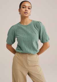 Top tricotat texturat verde cu mâneci scurte bufante, decolteu rotund și model în zig-zag, asortat cu pantaloni bej îmbrăcați pe măsură.