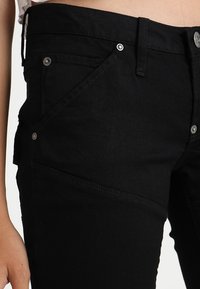 Černé džíny s úzkým střihem, pěti kapsami a stříbrnými kovovými detaily. Materiál má hladkou texturu s minimálním vyblednutím.