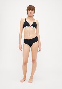 Bikini noir avec un bralette triangulaire doté d'une bande élastique blanche de marque. Le bas du bikini est également noir, avec une coupe classique.