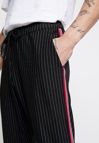 Zwarte broek met krijtstrepen en een trekkoord in de taille, voorzien van zijzakken en rode, witte en blauwe strepen langs de naden.