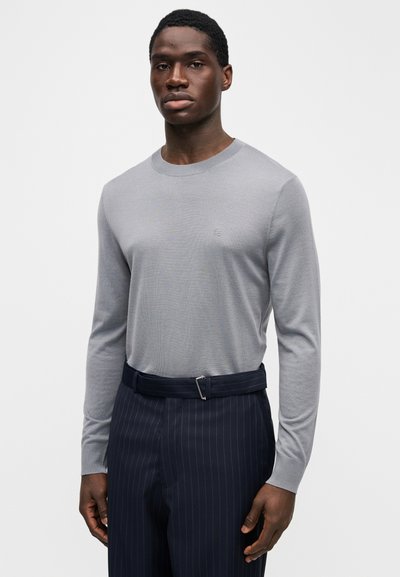 Chemise grise à manches longues en tissu fin tricoté, avec un col rond et un logo subtil, portée avec un pantalon bleu marine à fines rayures.