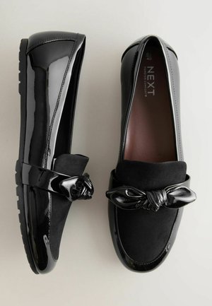 Mocasines negros que combinan materiales de charol y ante, con un nudo decorativo y diseño de punta redondeada, sobre una suela de goma texturizada.