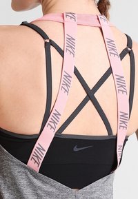 Black sports bra with pink adjustable straps. Crossed strap design features repeating "NIKE" branding. Gray fabric for additional coverage.