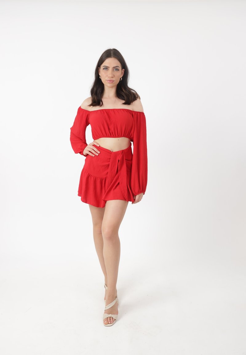 Frau steht mit der Hand auf der Hüfte, trägt ein rotes schulterfreies Crop-Top und einen passenden Rock, beige High Heels, vor einem schlichten weißen Hintergrund.