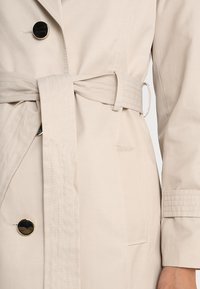 Beige trenchcoat med stora svarta knappar, bälte i midjan och strukturerade detaljer på ärmarna. Har framfickor och en slät tygfinish.