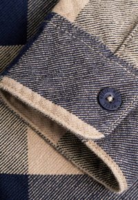 Puño de camisa de algodón con un patrón de cuadros en azul marino y beige, borde deshilachado y un botón azul marino con texto en relieve.