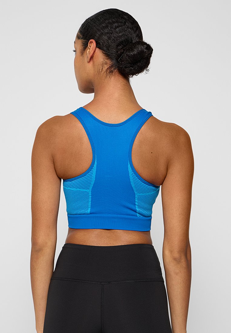 Mujer con el pelo recogido en un moño, vistiendo un sujetador deportivo azul de tirantes y leggings negros de cintura alta, vista desde atrás sobre un fondo sencillo.