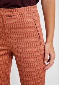 Orange patterned trousers with a diamond leaf design, featuring a side zipper pocket and smooth textured fabric.