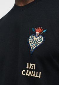 Μαύρο ύφασμα με κεντητό σχέδιο λουλουδιών σε σχήμα καρδιάς σε μπλε, πορτοκαλί και άσπρο πάνω από το κείμενο «JUST CAVALLI» σε κρέμ κλωστή.