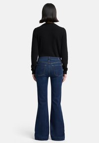 Dunkelblaue, ausgestellte Jeans mit hoher Taille, mit zwei Gesäßtaschen und kontrastierenden Nähten. Kombiniert mit einem schwarzen Langarmshirt.