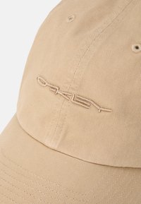 Beige színű baseball sapka texturált anyagból, elöl hímzett logóval és varrott részletekkel a peremen.