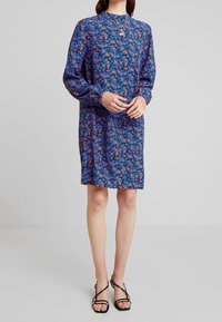 Femme portant une robe bleue à fleurs mi-longue avec manches longues et des sandales à talons hauts noires à brides, debout les mains jointes.