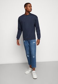 Sweatshirt de crewneck azul-marinho feito de algodão, combinado com jeans retos desgastados de corte claro e sapatilhas brancas com detalhes cinza.